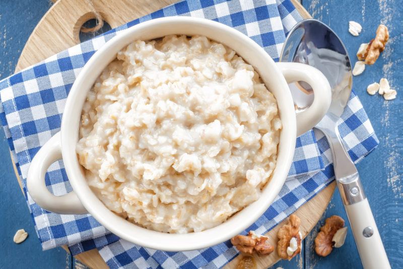 top view image of cooked oatmeal on a bowl placed on top of a blue gingham placemat with a spoon and loose nuts around it