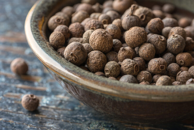 on a rustic looking wooden surface is a closeup image of a ceramic bowl full of allspice, with a couple of loose allspice