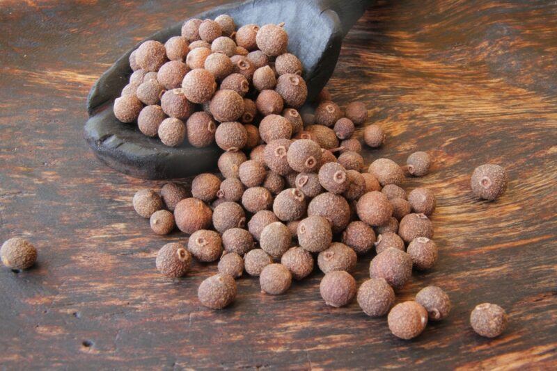 a closeup image of a small mound of allspice on a wooden surface with a wooden ladle also with allspice