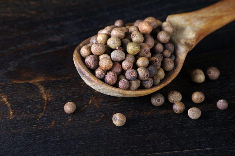 on a dark wooden surface is a wooden spoon full of allspices, beside it are a few loose allspice