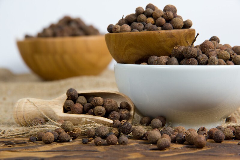 closeup image of wooden scoop with allspice with 3 different bowls at the back full of allspice