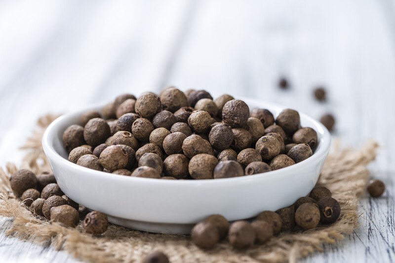 on a white wooden surface is a closeup image of a white dish full of allspice resting on a burlap sack with loose allspice around it