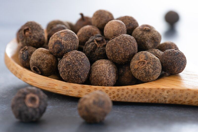 closeup image of a wooden spoon full of allspice, with loose allspice around it