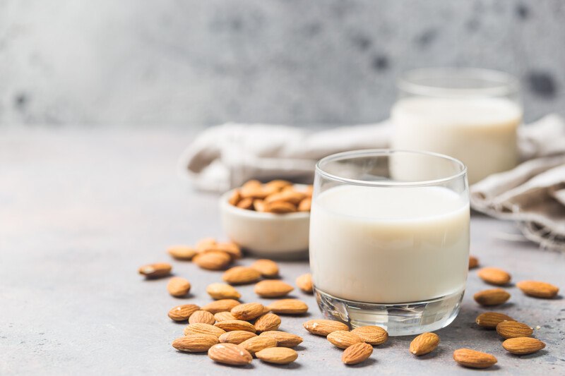 a couple of glasses with almond milk with a small white dish full of almonds and loose almonds around it