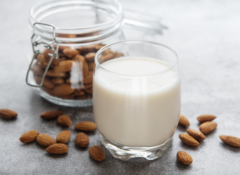 a glass of almond milk with a jar of almonds behind it and loose almonds around