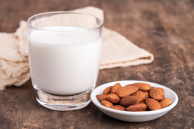 on a rustic looking wooden surface is a glass of almond milk with a white saucer full of almonds beside it and a beige table napkin behind it