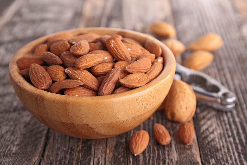 on an aged wooden surface is a wooden bowl full of almonds with loose almonds around it with a metal nutcracker