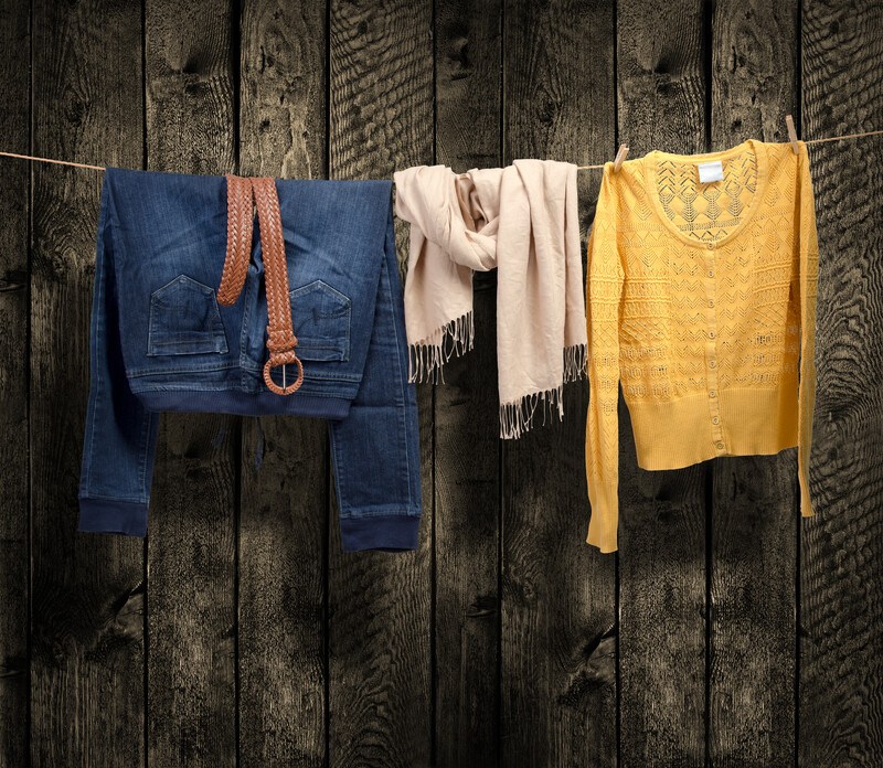 an outfit displayed on a clothesline with a barn wood wall in the background, from left to right a pair of jeans with leather belt draped over them, a cream colored scarf, and yellow button up sweater