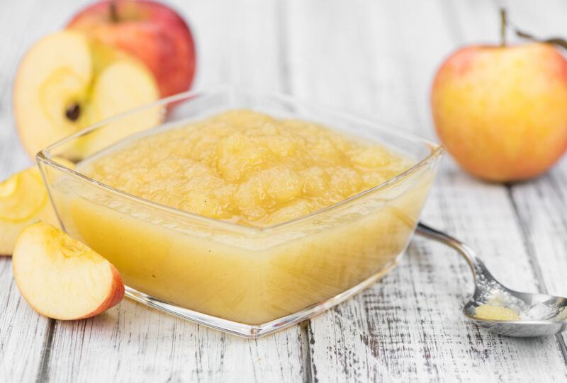 on a rustic looking white wooden surface is a square glass bowl with apple sauce, beside it is a silver spoon, around it are fresh apples some whole and some cut into slices