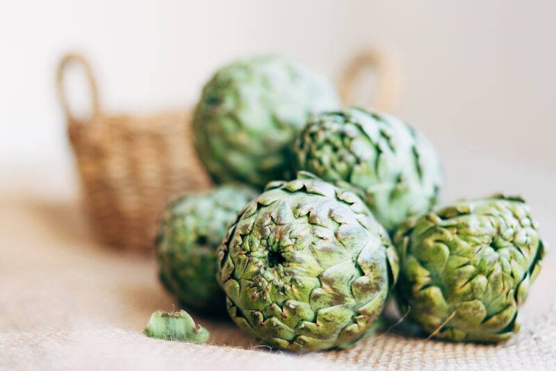 on a light brown cloth is a pile of fresh artichokes, a weaved basket can be seen at the back