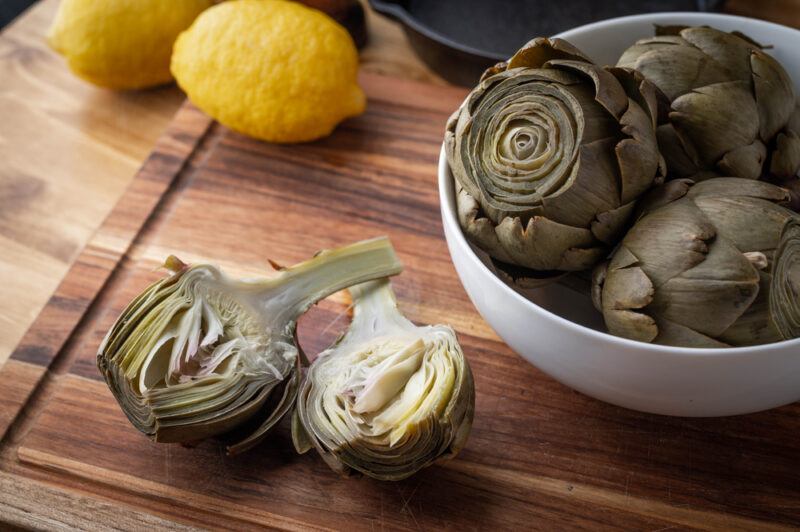 on a wooden surface is a halve artichokes with a white bowl full of artichokes, behind are a couple of whole lemons