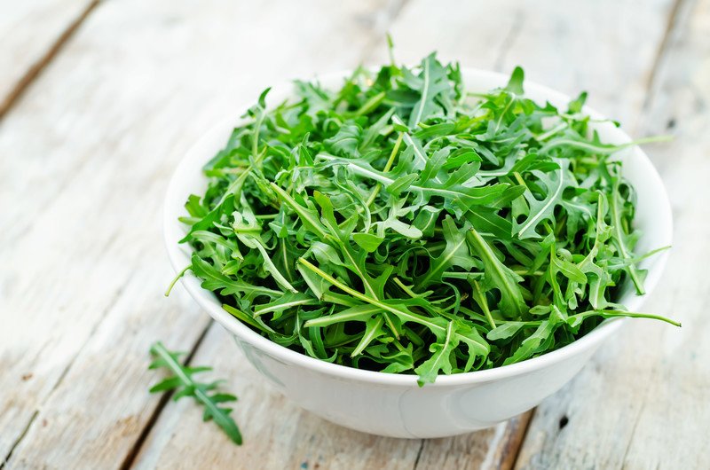 on an aged wooden surface is a white ceramic bowl full of arugula