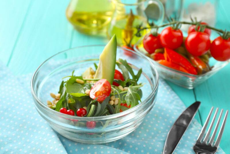 on a blue-green wooden surface is a clear glass bowl with arugula and cherry tomatoes with a slice of avocado on top, resting on top of a blue table napkin with small white dots, beside it is a silver knife and fork, at the back is a dish with a bunch of cherry tomatoes and a clear jar of olive oil