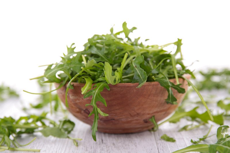 a closeup image of a brown wooden bowl full of arugula with some arugula scattered around it