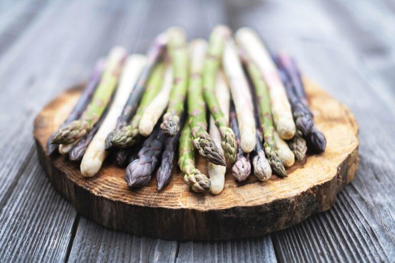 on a rustic looking wooden surface with a wooden round board with fresh green, white, and purple asparagus on it