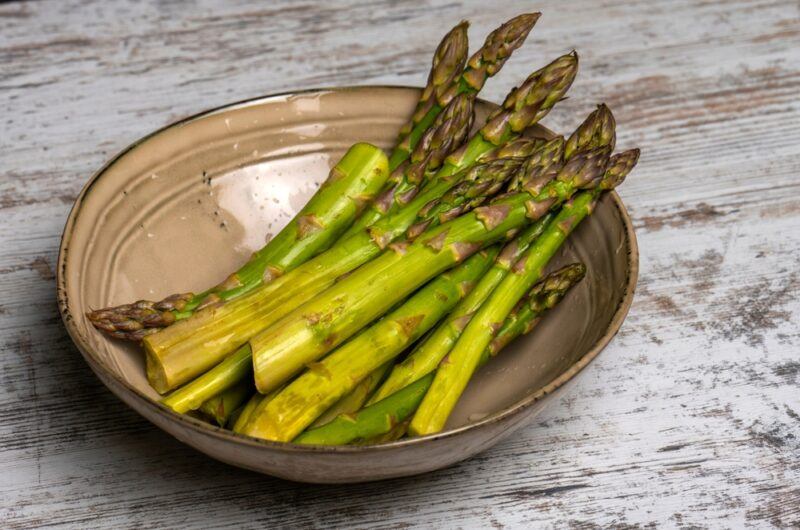 on a wooden surface is a tan-colored ceramic bowl with asparagus