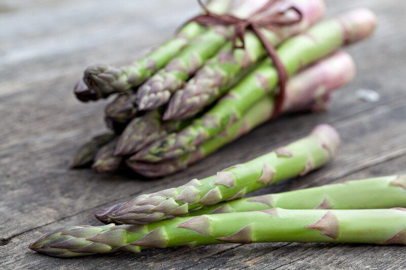 on a rustic wooden surface are three asparagus stems with a bundle of asparagus at the back