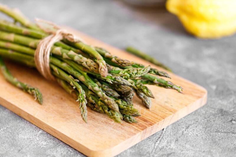 on a concrete surface is a wooden chopping board with a bunch of fresh asparagus, at the back is partial image of a lemon