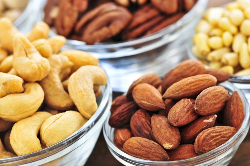 closeup image of different nuts in clear glass bowls, including cashew, almonds, and pecans.