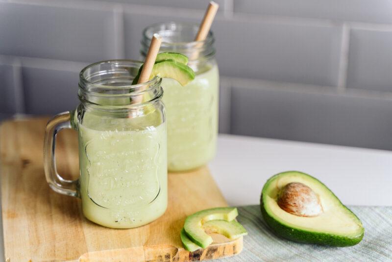 a couple of mason jars with handle full of avocado smoothie, resting on top a wooden chopping board, beside it is a sliced and halve fresh avocado