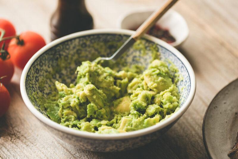 on a wooden surface is a white with intricate blue prints ceramic bowl with mashed avocado and a spoon with wooden handle, behind it is a bunch of cherry tomatoes, a pepper mill, and a small dish with spices