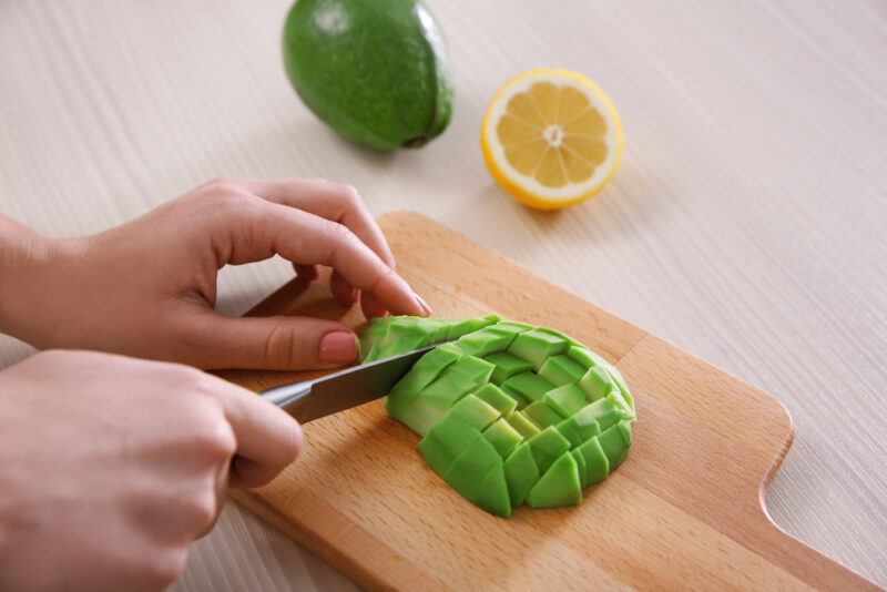 closeup image of a woman's hands slicing avocado on a wooden chopping board with a whole avocado and halve lemon beside it