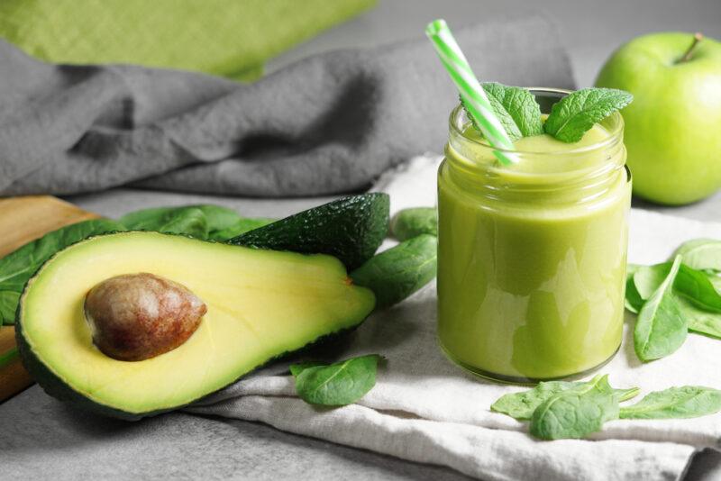 a mason jar with avocado and spinach smoothie, with green straw and sprig of herb on top, resting on a white table napkin with loose spinach leaves and halve avocado around it