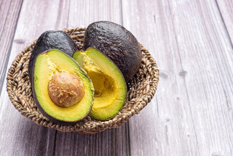 on rustic wooden surface is a brown round woven tray with a couple of whole avocados and one cut in halves