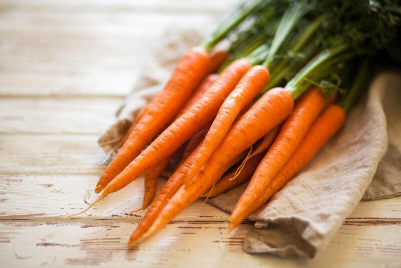 on a rustic white wooden surface are a bunch of baby carrots with leaves still attached resting on top of a brown table napkin