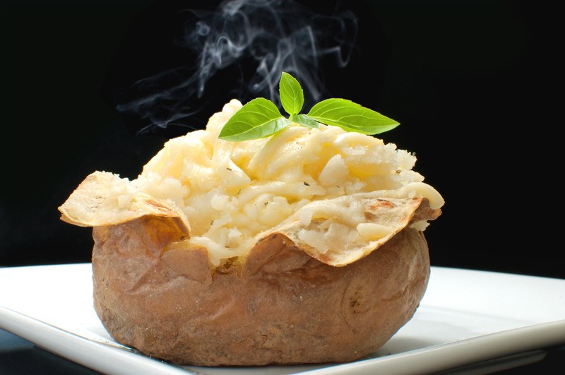 This photo shows a steaming baked potato with a sprig of green leaves on a white plate against a dark background.