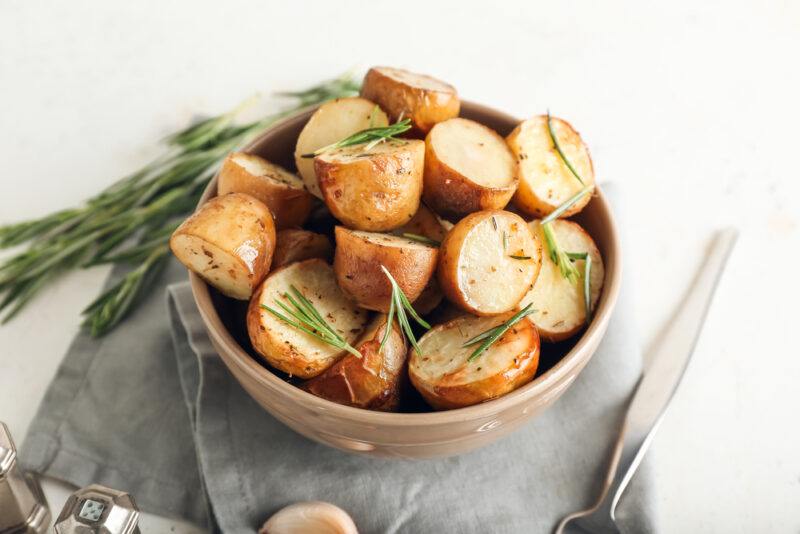 on a concrete surface is a light brown ceramic bowl full of baked potatoes with a sprinkling of rosemary herbs, resting on a light grey table napkin, beside it is a fresh sprigs of rosemary, silverware, a salt and pepper shaker