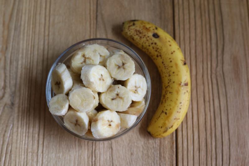 overhead shot of a wooden surface with a clear glass bowl full of sliced bananas, with a whole banana beside it