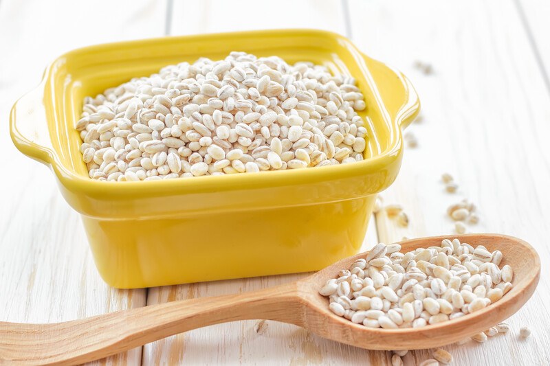 on a white wooden surface is a yellow square ceramic dish with barley, beside it is a wooden spoon with barley, with loose barley around it