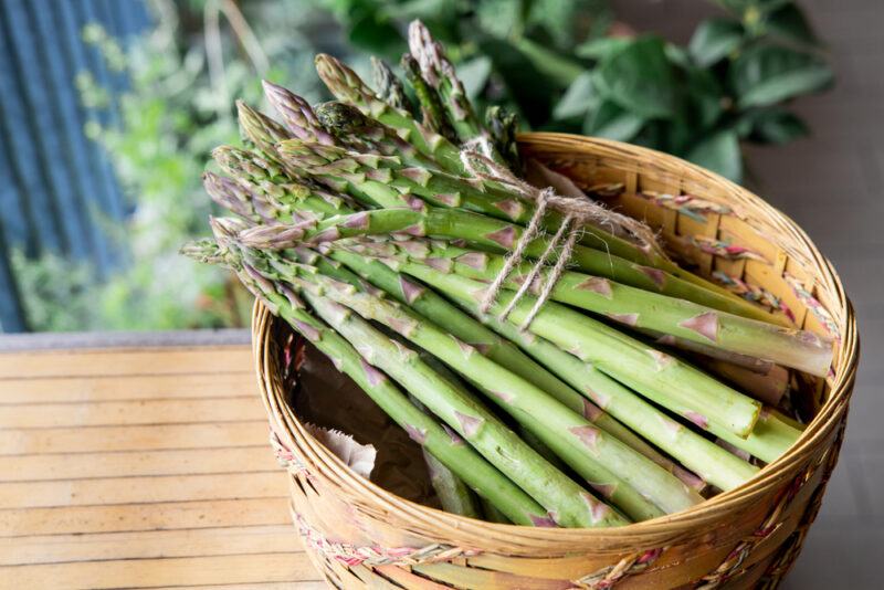 a basket of fresh asparagus on a wooden surface with leafy green vegetables at the back