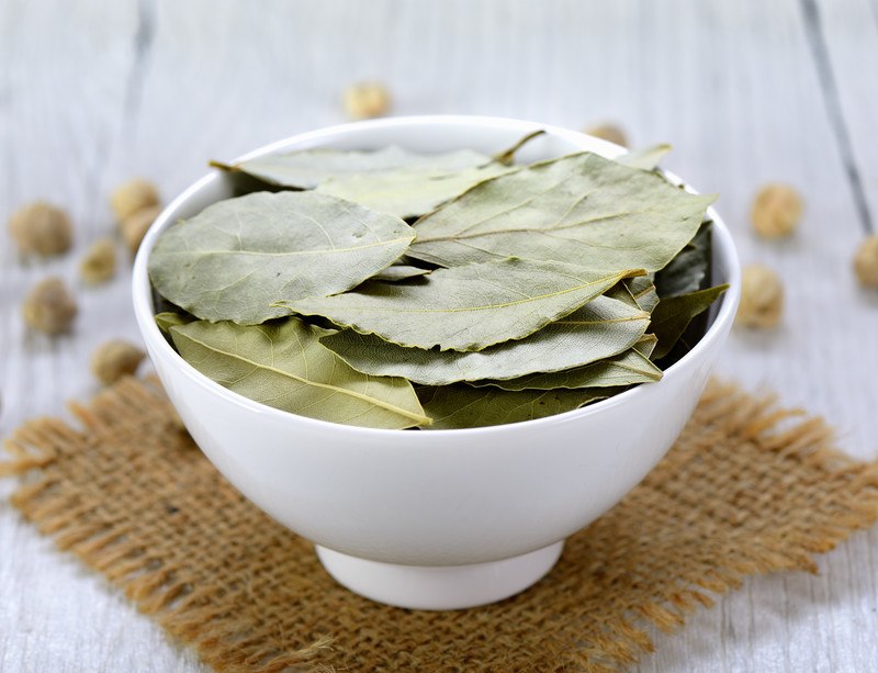 on a wooden white table is a white bowl full of dried bay leaves, with a piece of burlap underneath it, loose grains at the back