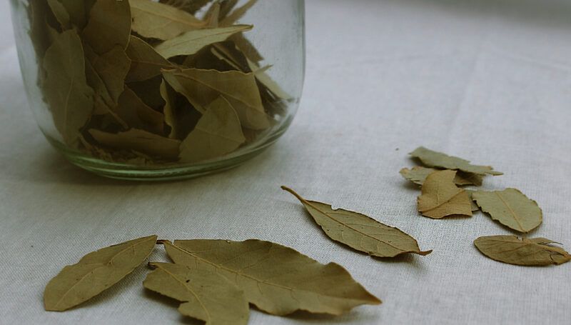 on a light-colored wooden surface are loose dried leaves beside a jar containing bay leaves as well