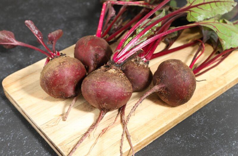 on a dark grey colored surface is a wooden chopping board with a bunch of fresh beets