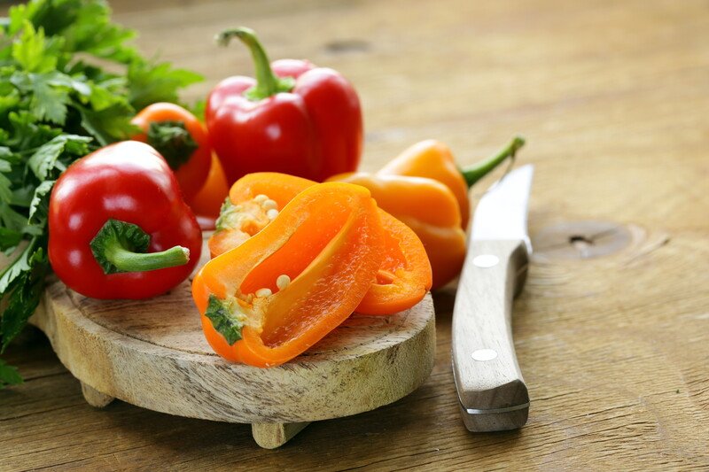 on a wooden chopping board is a yellow and red bell peppers with a knife beside it and fresh herbs at the back