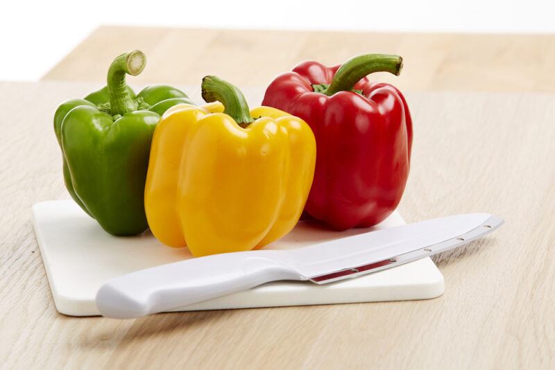 on a light brown wooden surface are yellow, green, and red bell peppers on a white chopping board and white knife