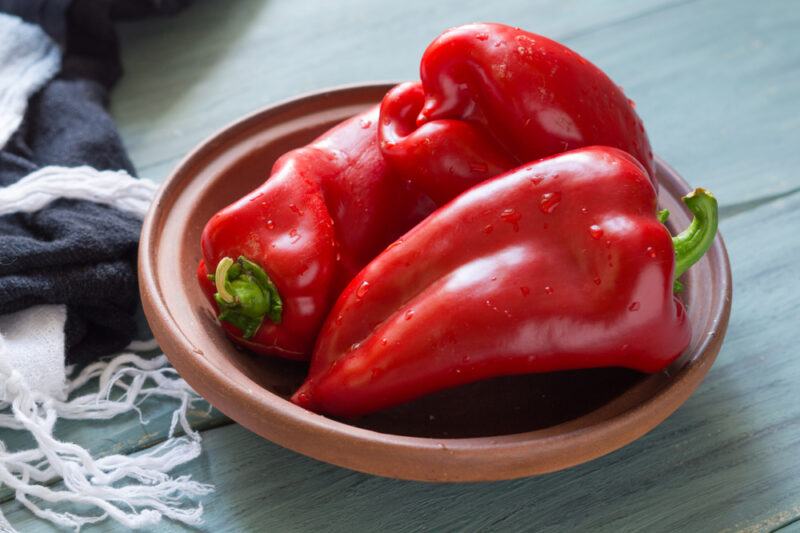 on a wooden surface is a clay shallow bowl with three red bell peppers