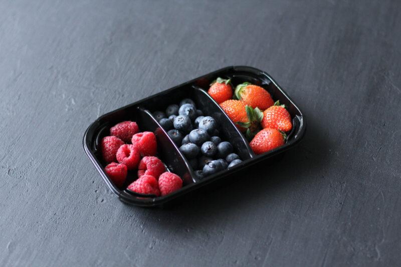 on a dark grey wooden surface is a black plastic container with berries including raspberries, blueberries and strawberries