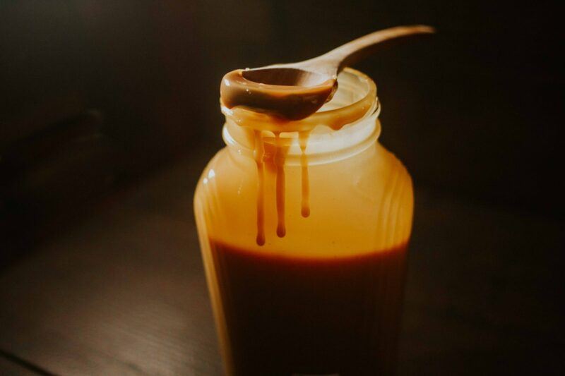 a glass jar filled with caramel and a wooden spoon with caramel dripping off it to represent the bests caramel vodka