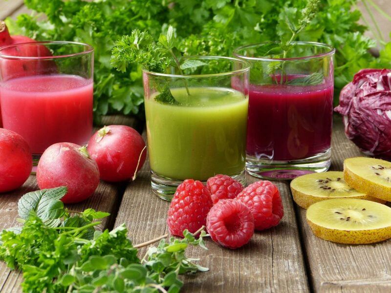 three glasses of colorful fruit juice surrounded by fruits and greens to represent the best fruit juice brands