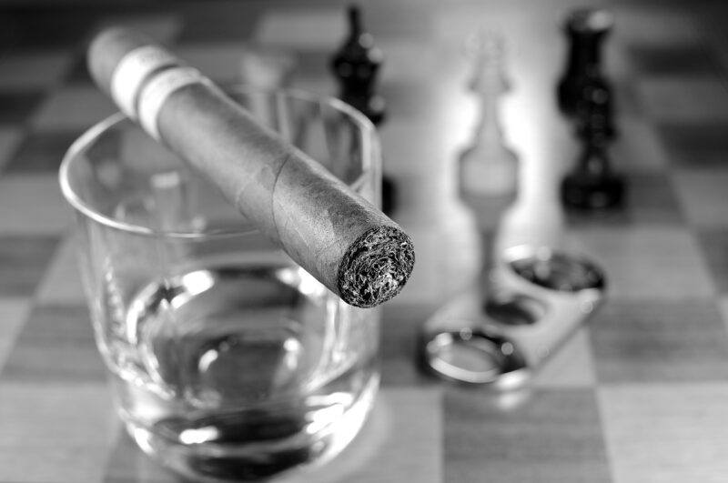 best-gifts-for-whiskey-lovers | Food For Net a black and white photo of a small glass of whiskey with a cigar across the top with a blurred chess board in the background to represent the best gifts for whiskey lovers