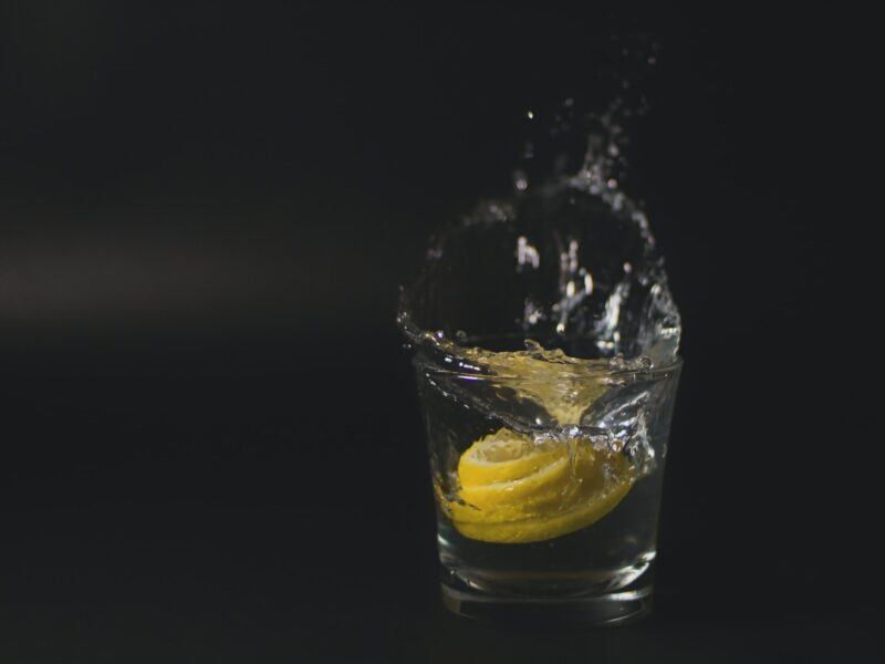 a shot glass with vodka splashing out of it and a circle of lemon in the glass on a black background to represent the best lemon vodka