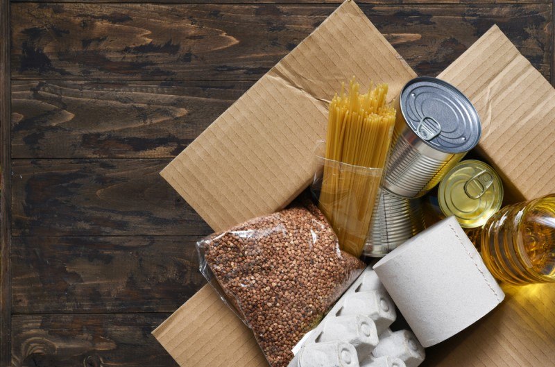 A cardboard box containing pasta, several cans of food, a bottle of cooking oil, a bag of brown beans, and a roll of toilet paper rests on a wooden surface, representing the best non perishable foods for emergency use.