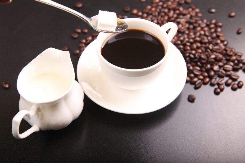 best-plant-based-sweetener- | Food For Net a cup of coffee next to some creamer, surrounded by coffee beans and a sugar cube in a spoon to represent the best plant-based sweetener