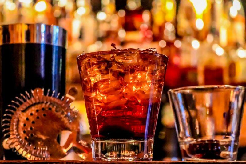 a full glass of rum on ice with a bright, blurred bar scene behind it to represent the best rum to drink neat 