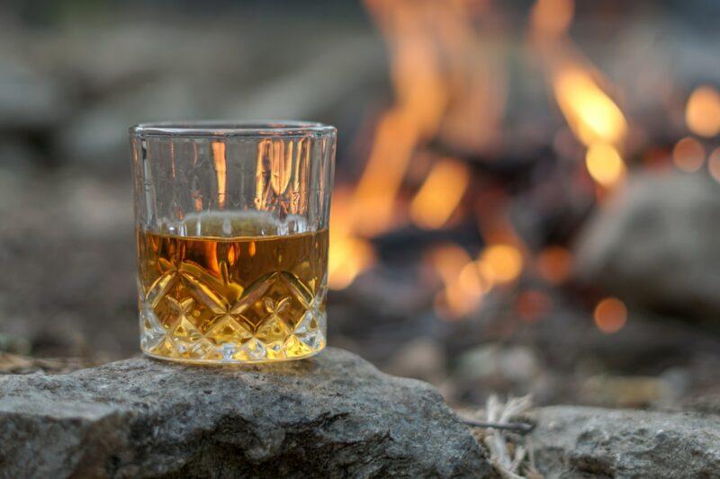 a glass of straight whiskey sitting on a rock with a blurred campfire in the background to represent the best wheat whiskey