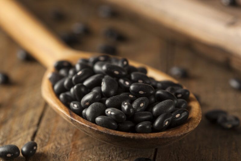 closeup image of a wooden ladle full of black beans resting on a wooden surface with loose black beans